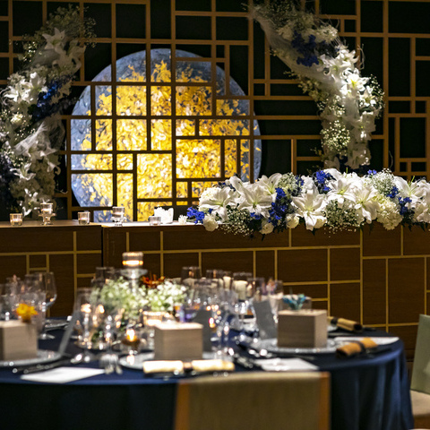 おふたりの披露宴会場「アルブル」は、黒×ゴールドを基調としたシックなバンケット。 全体的に白い装花を中心にポイントで青を入れたコーディネートに。 月のまわりの装花がより華やかに演出します。